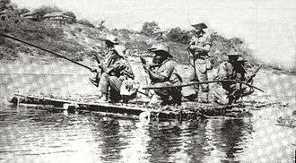 A raft on a river. A man with a rifle stands in the centre, surrounded by other men kneeling on one knee