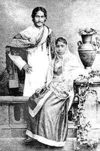 Black-and-white photograph of a finely dressed man and woman: the man, smiling, stands akimbo behind a settle with a shawl draped over his shoulders and in Bengali formal wear. The woman, seated on the settle, is in elaborate dress and shawl; she leans against a carved table supporting a vase and flowing leaves.