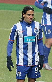 Man with long dark hair held with a headband, wearing a football kit composed of a shirt with vertical blue-and-white stripes and blue shorts. He also wears a blue sleeved undershirt and winter gloves.