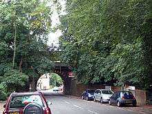 A street with several parked cars and large trees is crossed by a railway bridge