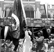 A man in military uniform raising a flag up a pole. Behind him are other uniformed men and others wearing traditional, civilian dress