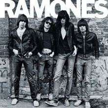 Four men standing against a graffiti-covered wall. Each man has a black leather coat, blue jeans, and brown hair. At the top of the black-and-white image, "RAMONES" is spelled out in all caps.