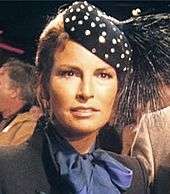 Welch in blue scarf and high-collared gray jacket, with polka-dot feathered cap.