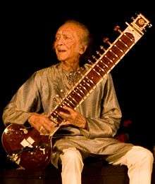 An elderly man is seated at the centre of the image, holding a sitar.