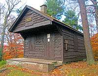 A small maroon-colored wooden cabin with its roof overhanging the front porch. On that section is a sign that says, in gold lettering, "Red Hill, 2,990 feet". Behind it are woods, partly green and partly in fall color