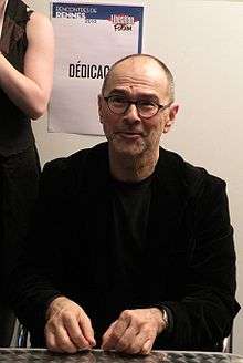 Head and shoulders portrait of sitting 60-year-old man in rimless glasses, black suit, and black tee-shirt, with both hands lying on a table.