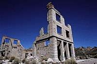 Ruins of a three-story masonry building rise into a cloudless, dark blue sky. The building is roofless, and large sections of it walls are missing. Masonry rubble lies about the building, which has openings for doors and windows but no glass or wood. Another ruin is nearby, and barren hills are visible in the distance.