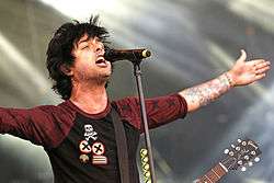 A man with black hair in a red and black baseball-style T-shirt sings into a microphone with his arms outstretched.