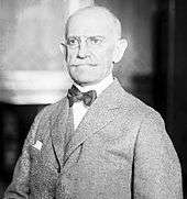 A bald man with glasses wearing a gray jacket and vest, white shirt, and black bowtie