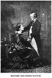 A couple is shown. On the left is a tall woman of about 30. She wears a voluminous dress and is sitting sideways in an upright chair, facing and looking up into the eyes of the man who is on the right. He is about 60, quite short, balding at the temples. He is dressed in a suit with tailcoat and wears a cravat. He faces and looks down at the woman. His hand rests on the back of the chair.