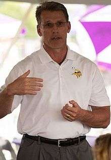 Candid photograph of Spielman wearing grey slacks and a white polo-style shirt bearing a Minnesota Vikings logo and gesturing with his hands