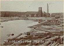 Sepia-tone photo of a pond surrounded by large logs. At the far end of the pond is a large building with a square tower and two smokestacks. Label is "T&T L CO MILL POND RICKETTS PA 1903" (i.e. Trexler and Turrell Lumber Company Mill Pond ...)