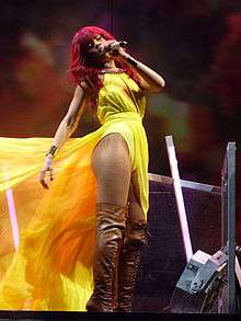 A woman with bright red long hair is singing. She wears a yellow dress with a long train, which is moved by the wind. Also the woman wears tights and brown boots.