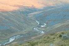 Ribbon of river in shadow reflecting the sky, bending back and forth through a valley between moorland hills of grey scree and brown and green vegitation