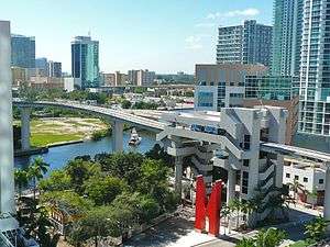 A small metro train on an elevated rail line crosses over a wide river, surrounded by high-rise buildings. An elevated metro station with a large "M" sculpture is in the foreground.