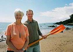 Jeanette Fitzsimons wearing an orange T-shirt, in front of Rob Hamil in a green shirt and holding an orange rowing oar. They are on the seashore.
