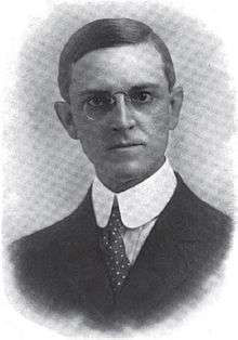 A man with dark hair wearing glasses, a high-collared white shirt, polka-dotted tie, and black jacket