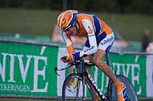 A road racing cyclist in an orange, blue, and white jersey and a matching helmet sits crouched low on his bicycle in aerodynamic position, with a grimace on his face.