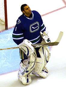 An unmasked ice hockey goaltender. His hair is slicked back and he is looking upwards. He wears white goaltending pads and a blue jersey with a logo of a stylized hockey stick.