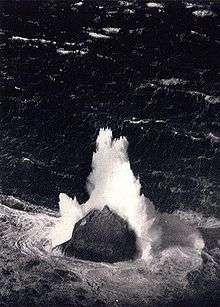 A back and white aerial image of an isolated and steep-sided rock in the midst of a stormy sea. A large wave has broken on the rock and sent fountains of white water high into the air.