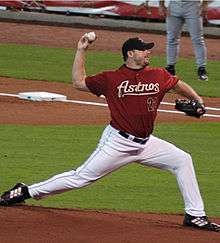 A man in white pants and a maroon baseball jersey with "ASTROS 22" on the chest pitches a baseball with his right hand.