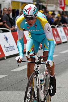 A road racing cyclist in a blue and yellow skinsuit with white trim, riding up out of the saddle. Spectators are visible in the background behind barricades