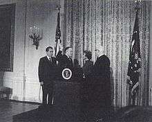 A formal-looking room with flags and drapery; in it, a middle-aged man, another middle-aged man with his right hand raised, a middle-aged woman, and an older man, are all beside a podium