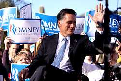 Mitt Romney sitting outdoors during daytime, with crowd behind him holding up blue and white "Romney" signs