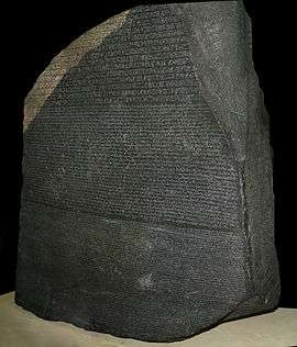A large, ancient, black-colored stone block with written inscriptions covering one side of its surface, with pieces clearly broken off with now missing text