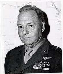 A black-and-white photo of Ross E. Rowell, a white male, in his Marine Corps uniform. There is a map in the background behind him, his ribbons are on the left breast side of his uniform directly above the pocket and he is wearing 2 star general rank insignia on each shoulder of his uniform. Directly above his ribbons he is wearing his Naval Aviator wings.