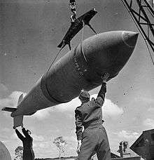 Black and white photo of a large bomb being hoisted. Two men wearing military uniforms are standing below the bomb, and steadying it with their hands.