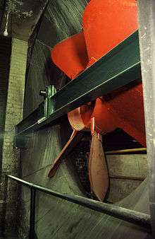 Large red six-bladed fan, fitted to a steel girder, enclosed inside a concrete and brick funnel.