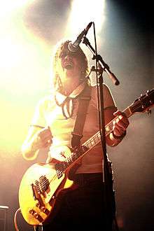 A Caucasian female with shoulder-length brown hair wearing a white shirt and dark pants sings into a microphone while strumming a yellow electric guitar.