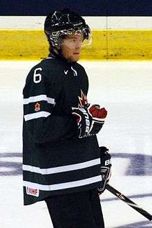 A teenage ice hockey player standing relaxed on the ice. He wears a green jersey with white trim and a black, visored helmet.
