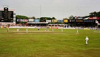 View of a cricket stadium. Players in Test cricket outfits can also be seen.