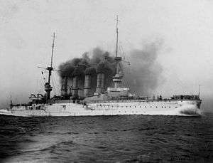 A large warship steams at full speed; black smoke billows from its four funnels
