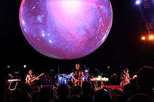 The Smashing Pumpkins perform on a stage with a large orb with projections. From left to right: Nicole Fiorentino, Billy Corgan, and Jeff Schroeder.
