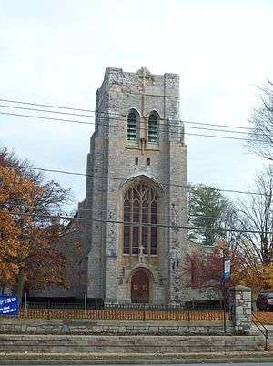A stone church tower with a level top, gently arched pointed windows and a small wooden door in the base. In front of it are telephone lines and a set of steps.