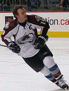 A hockey player with short, brown hair looks to his left as he skates. He is in a white and burgundy uniform with a stylized A logo on his chest.
