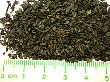 A pile of brownish-dark green dried leaf pieces, with a ruler for measurement. The pieces are generally round with diameters of 1 to 4 mm. Some pieces are rectangular, about 1 to 3 mm wide and 3 to 8 mm long.