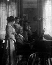 Photograph of Mark Twain playing piano, with his daughter Clara and her friend behind him