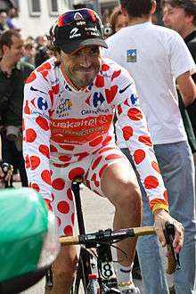 A road racing cyclist of about thirty, wearing a jersey and bike shorts adorned with decorative polka dots, a black baseball cap, and sunglasses on the cap. He has a smirk on his face, and various people stand behind him.