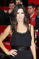 A portrait of Sandra Bullock wearing black dress, with paparazzi standing in the background.