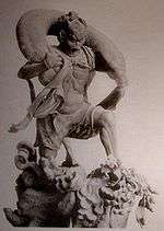 Fūjin. Three-quarter view of a statue. His left leg is bend as if climbing stairs and he is carrying a long bag-shaped object which goes from one shoulder to the other around the back of his head. Black and white photograph.