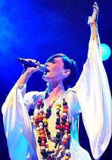A 34-year-old woman is shown in left profile, she is singing into a microphone in her right hand, with her head titled back and looking forward. Her left arm is raised in a white robe with wide sleeves. She wears a multi-stranded necklace with numerous beads of various sizes and colours.