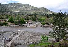 A muddy river flows through a gap between the ruins of a dam on either shore. A wooded hill or mountain rises in the background.