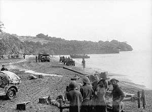 Soldiers wearing steel helmets on a beach, manning an antiaircraft gun. In the background are jeeps and trucks, and an LST.