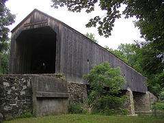 The front entrance and right side of an unpainted covered bridge supported by stone piers