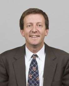 A portrait shot of Scott S. Harris, looking straight ahead. He has short brown hair and is wearing a charcoal blazer with a blue patterned tie over a white collared shirt.