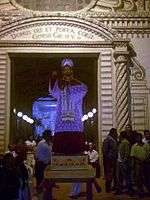 A statue of St. Peter placed on a pedestal in front of an entrance to the church. The facade of the church and inside is lit up and people are standing around the statue.